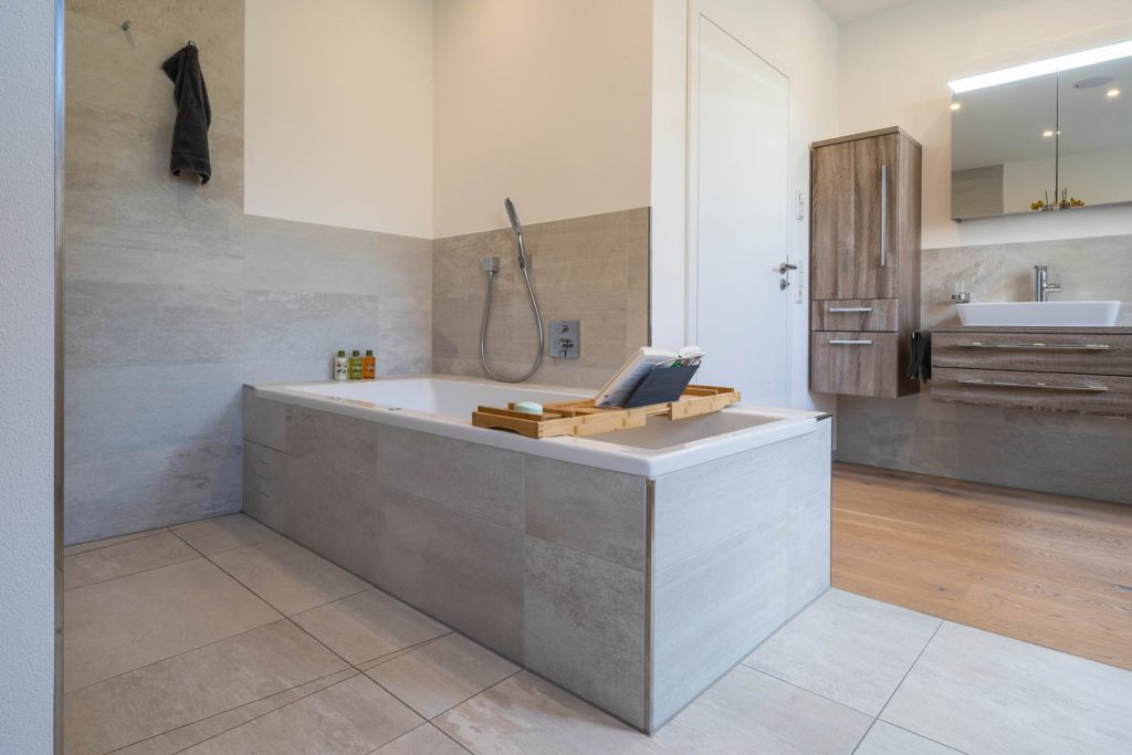 Badewanne mit Fliesenverkleidung in Steinoptik, rechts Holzboden im Waschbereich
