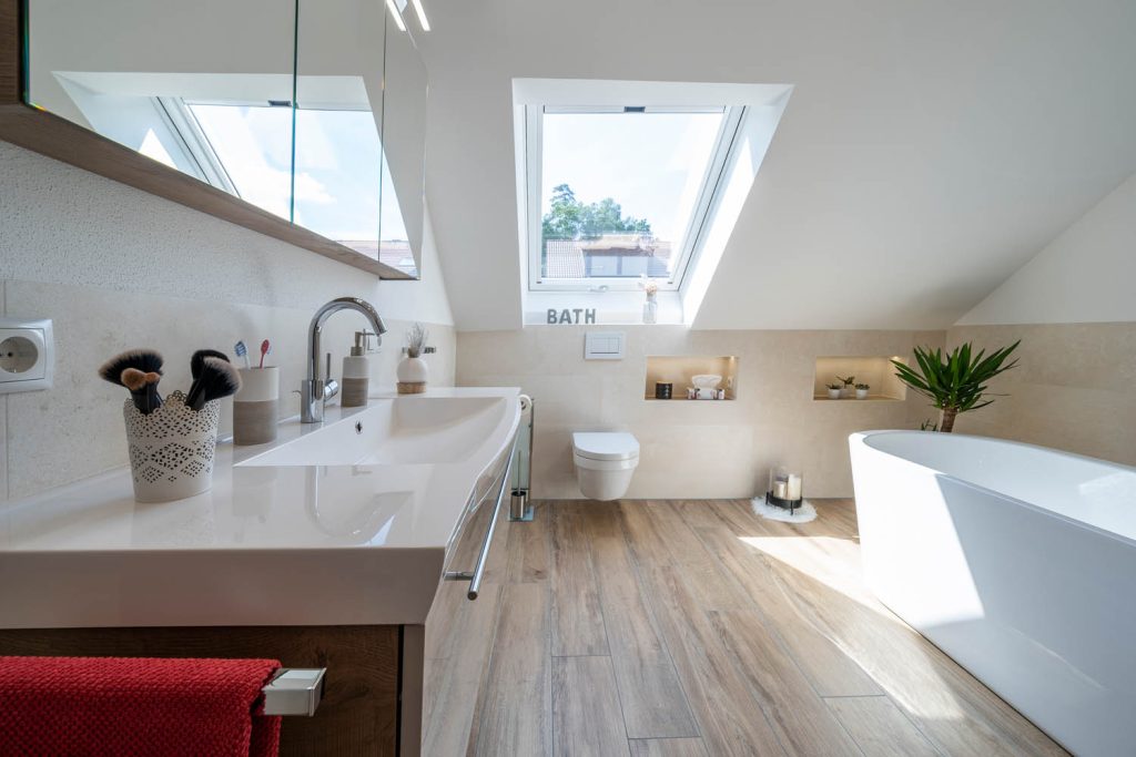 Waschtisch unter dem Dachfenster, dahinter Wand-WC und freistehende Badewanne