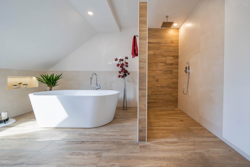 Freistehende Badewanne vor beleuchteten Wandnischen, rechts die begehbare Dusche