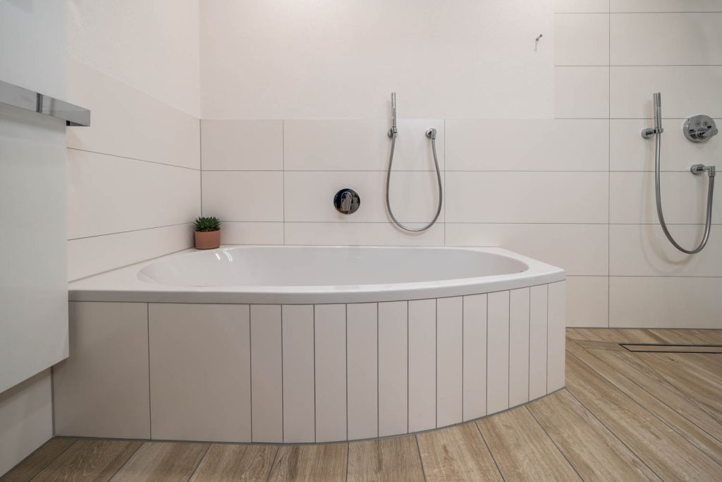 Eckbadewanne mit weißer Verkleidung und Handbrause vor hellen Wandfliesen