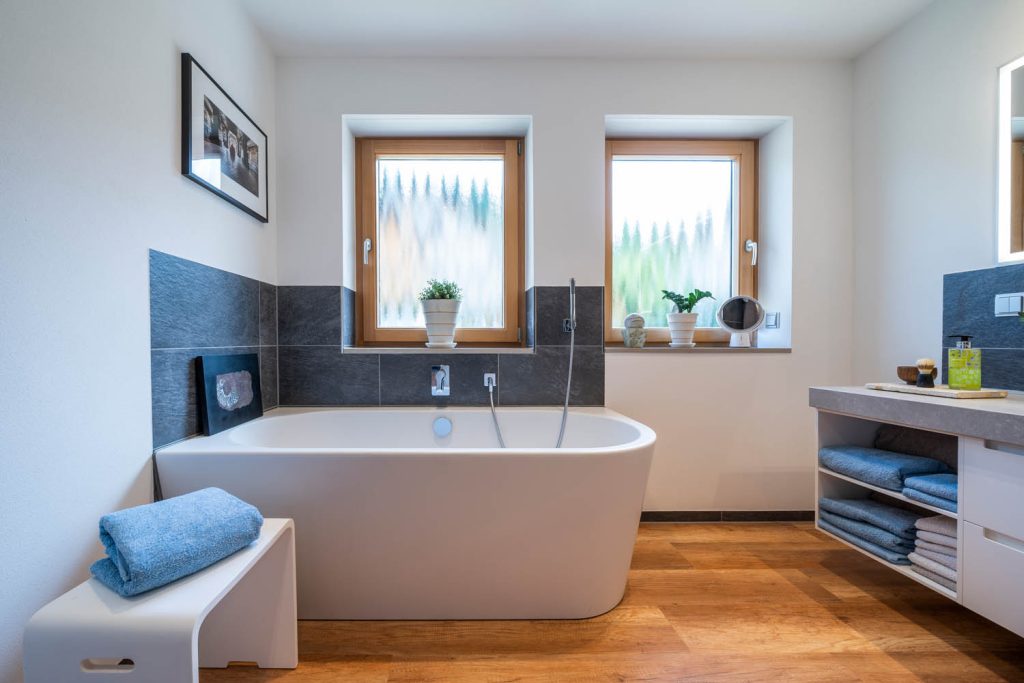 Freistehende Wanne unter zwei Fenstern vor dunklem Fliesensockel, links ein Hocker