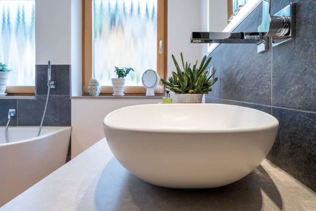 Rundes Aufsatzbecken mit Wandauslauf, im Hintergrund Fenster und Wanne