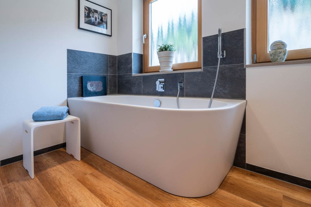 Freistehende Wanne vor dunklem Fliesensockel mit Hocker und Handtuch