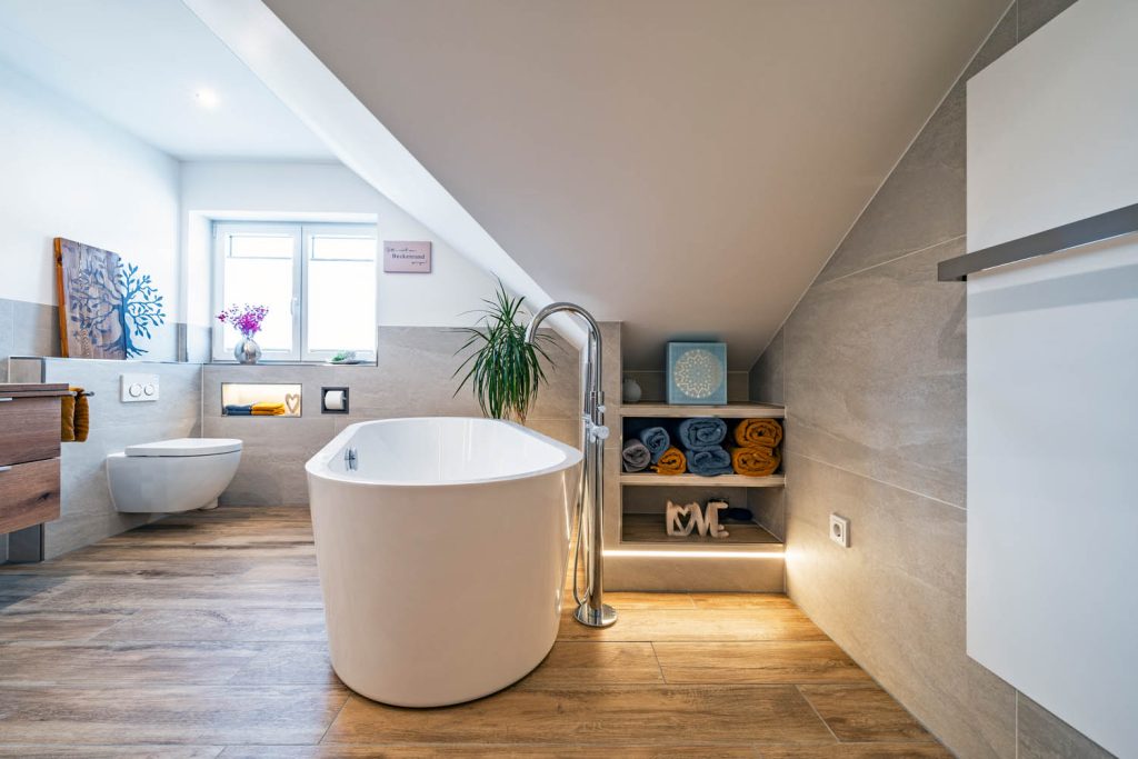 Freistehende Wanne mit Standarmatur vor beleuchtetem Regal mit Handtüchern