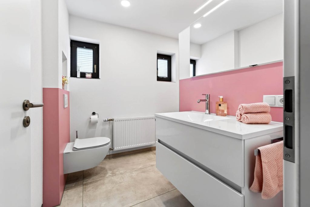 Blick durch die Tür ins Bad: weißer Waschtisch vor rosé Wandfläche, Wand-WC und Heizkörper unter zwei Fenstern