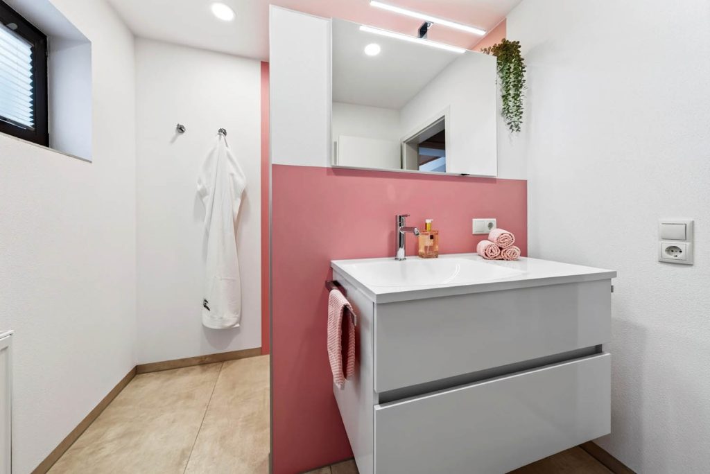 Waschtisch mit weißem Unterschrank und rosa Handtuch vor rosé Wand, daneben ein Haken mit Bademantel