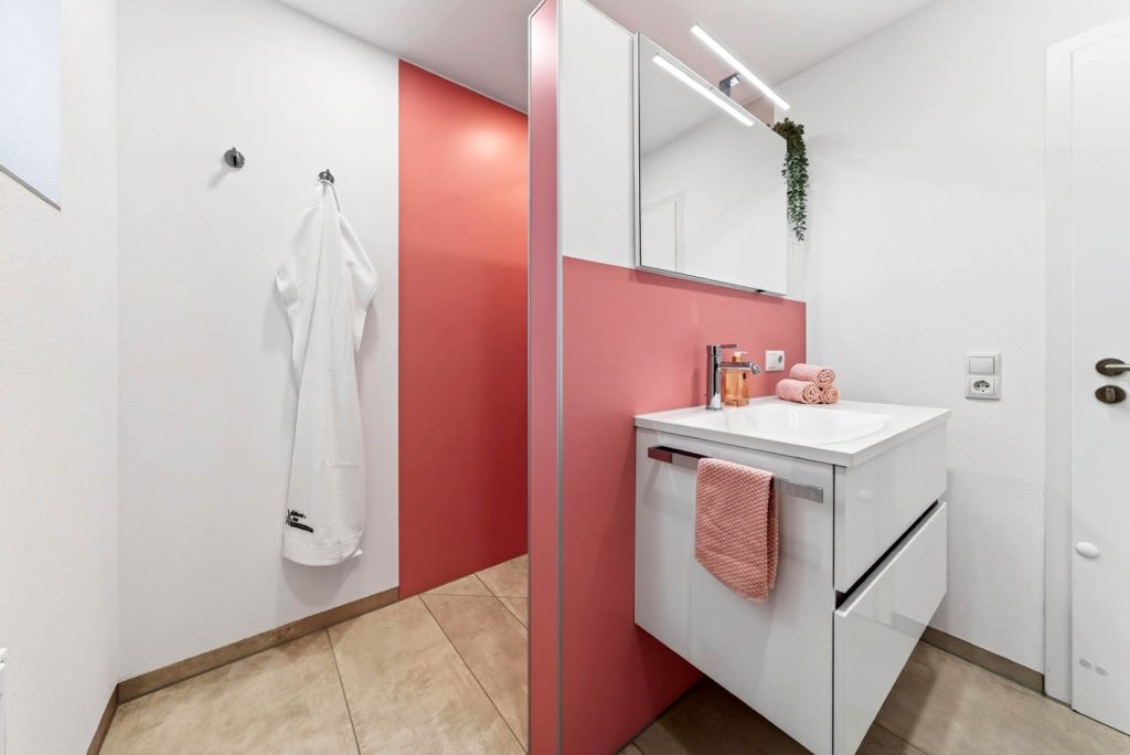 Rosé Wandscheibe zwischen Waschbereich und Dusche, an der weißen Wand hängt ein Bademantel