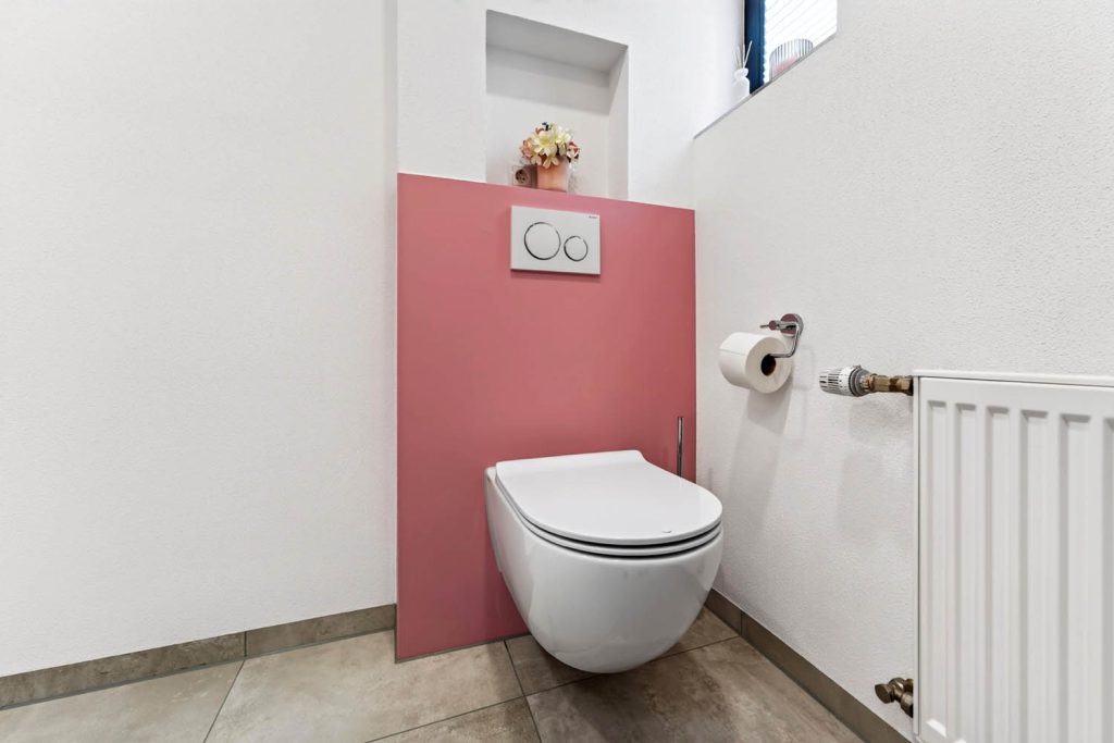 Wandhängendes WC vor rosé Vorwand mit Ablagenische, daneben Papierhalter und Heizkörper