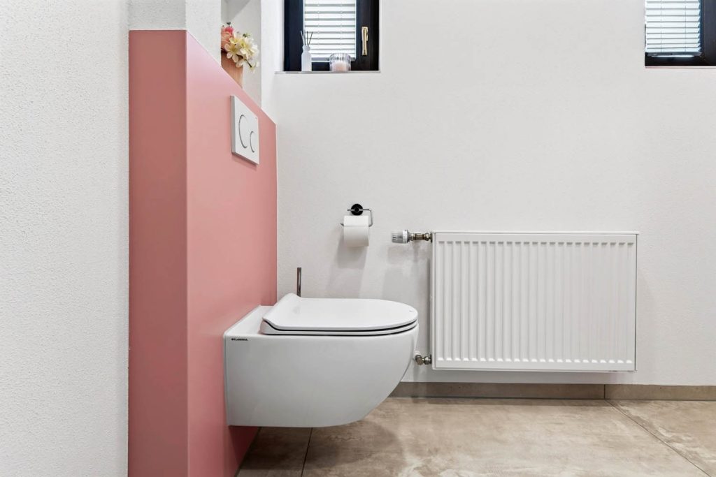 Wand-WC neben rosé Vorwand, an der weißen Wand Heizkörper und Papierhalter