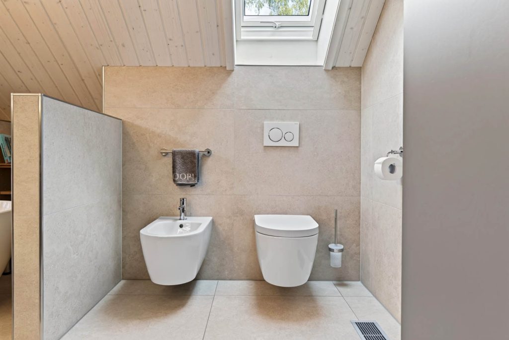 Wand-WC und Bidet vor großformatigen Fliesen in heller Steinoptik unter dem Dachfenster