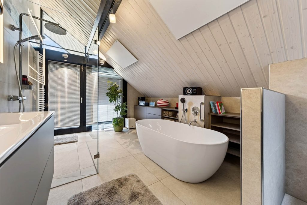 Freistehende Wanne vor einer Vorwand mit Wannenarmatur, links die verglaste Dusche