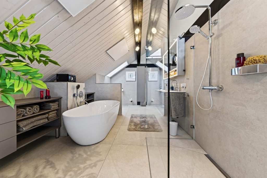 Badewanne und Regalwand unter der Dachschräge, rechts die bodengleiche Dusche mit Glaswand