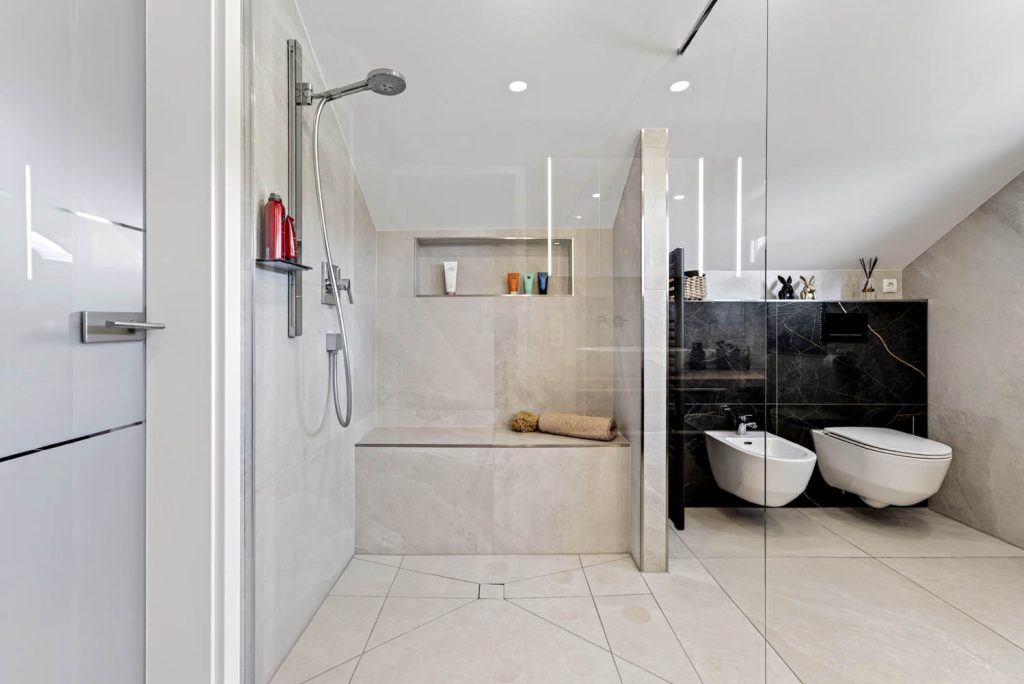 Blick in die Dusche mit Glaswand und Sitzbank, rechts Bidet und WC vor dunklen Fliesen