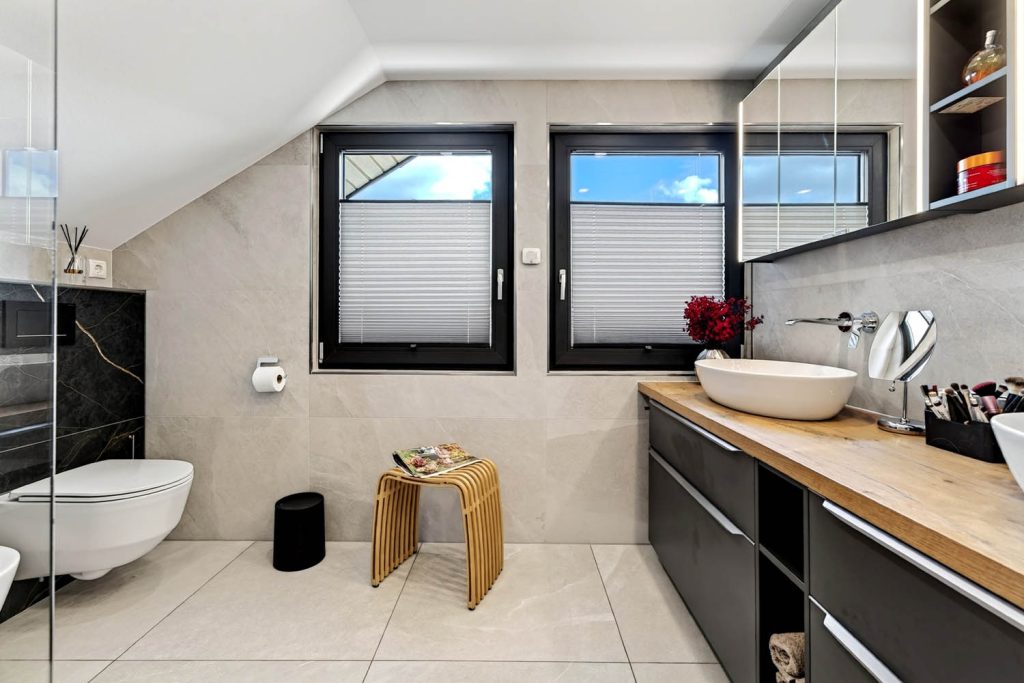 Raumansicht mit Wand-WC, Hocker aus Holz und Waschtisch unter zwei Fenstern