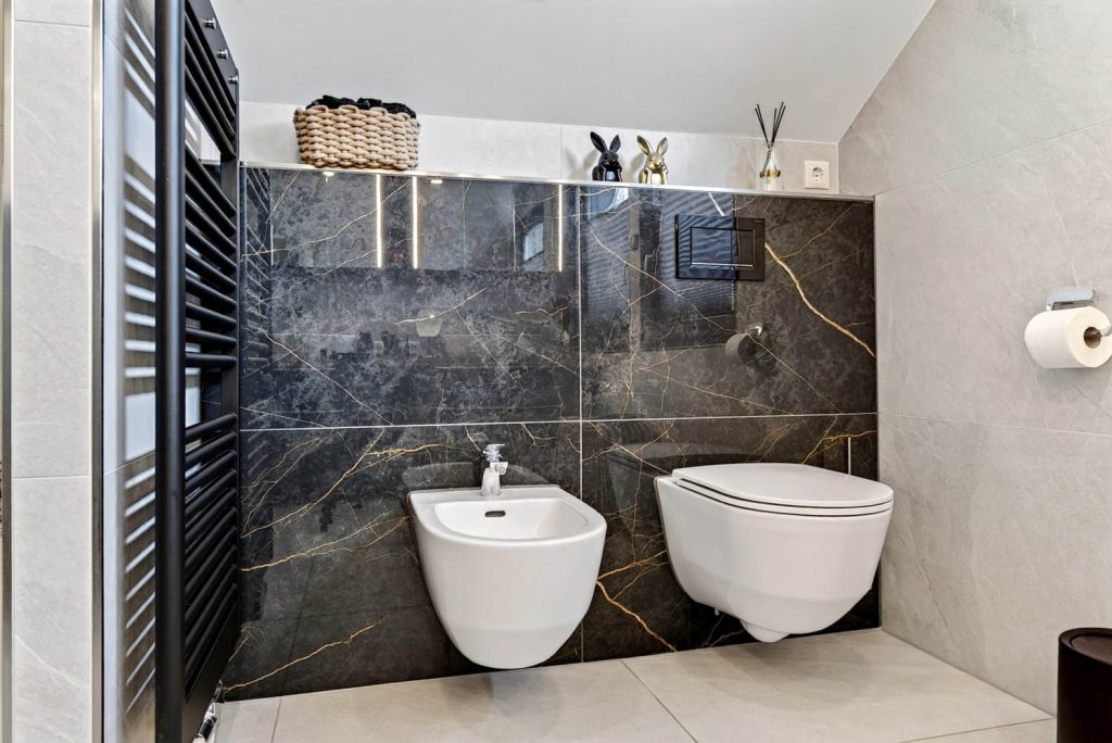 Bidet und WC vor dunkler Marmorwand mit Ablage, links ein schwarzer Handtuchheizkörper