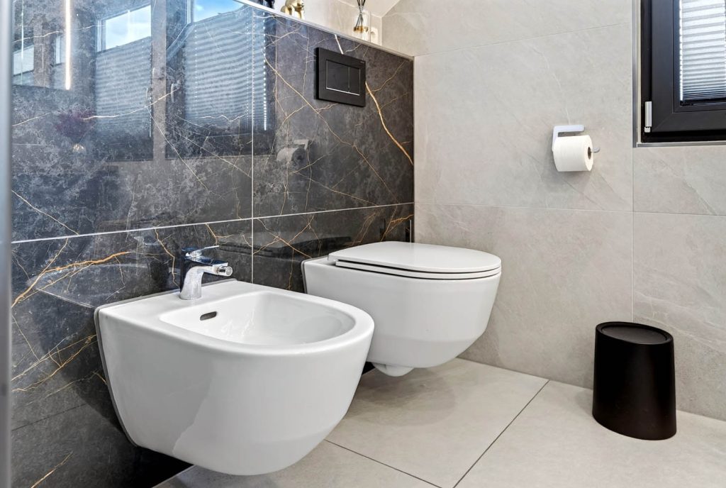Detail von Bidet und Wand-WC vor dunklen Fliesen mit goldfarbener Maserung
