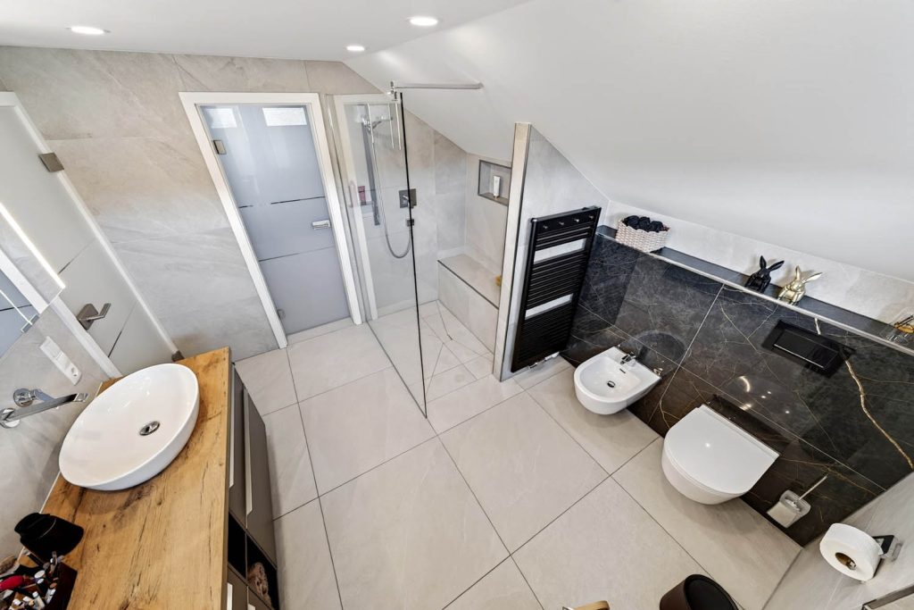 Weitwinkel mit Dusche, dunkler Marmorwand im WC-Bereich und Waschtisch im Vordergrund