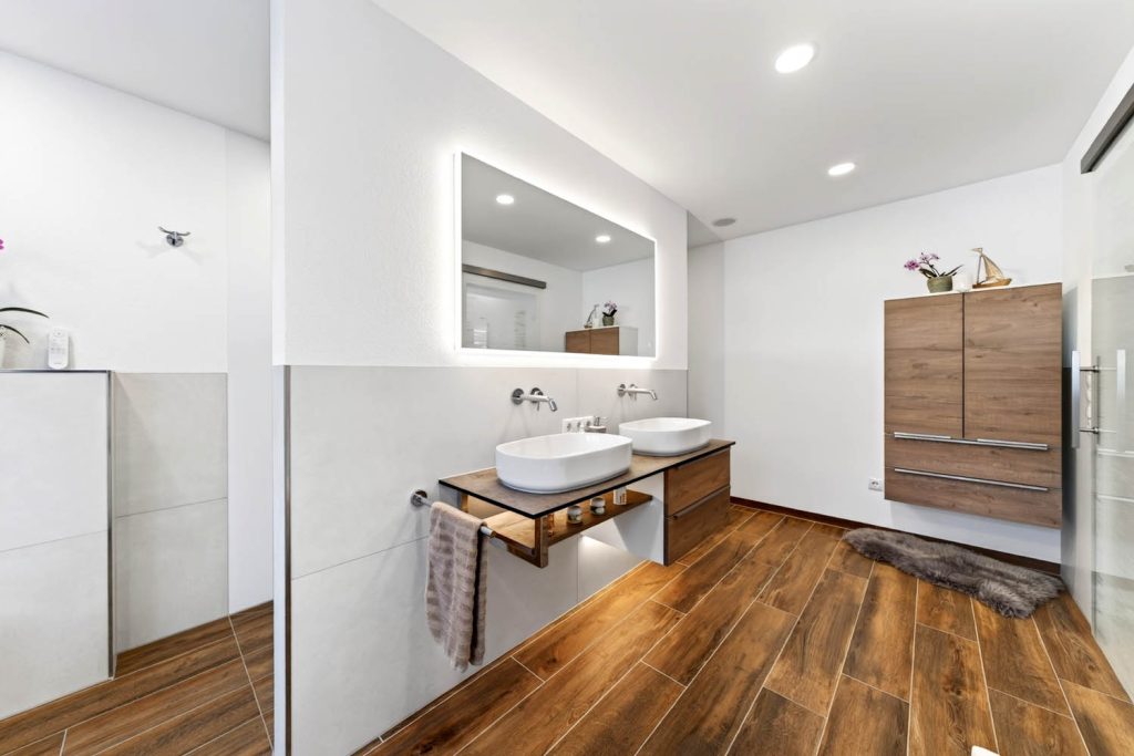 Waschtisch an der Vorwand mit beleuchtetem Spiegel, rechts ein Hängeschrank in Holzoptik