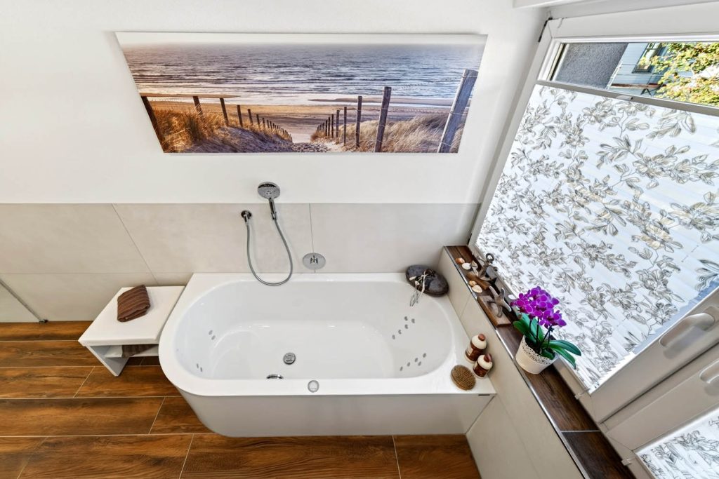 Eckbadewanne mit Sitzbank unter einem Strandbild, rechts das Fenster