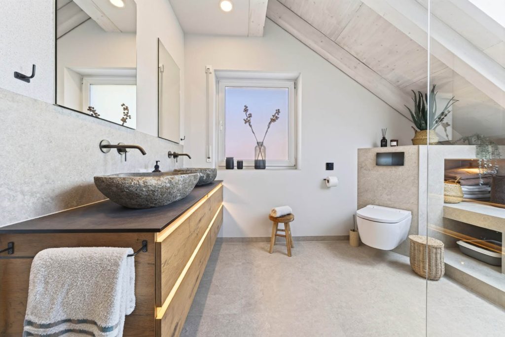 Waschtisch mit Natursteinbecken und Handtuch, dahinter WC-Vorwand und Badewanne