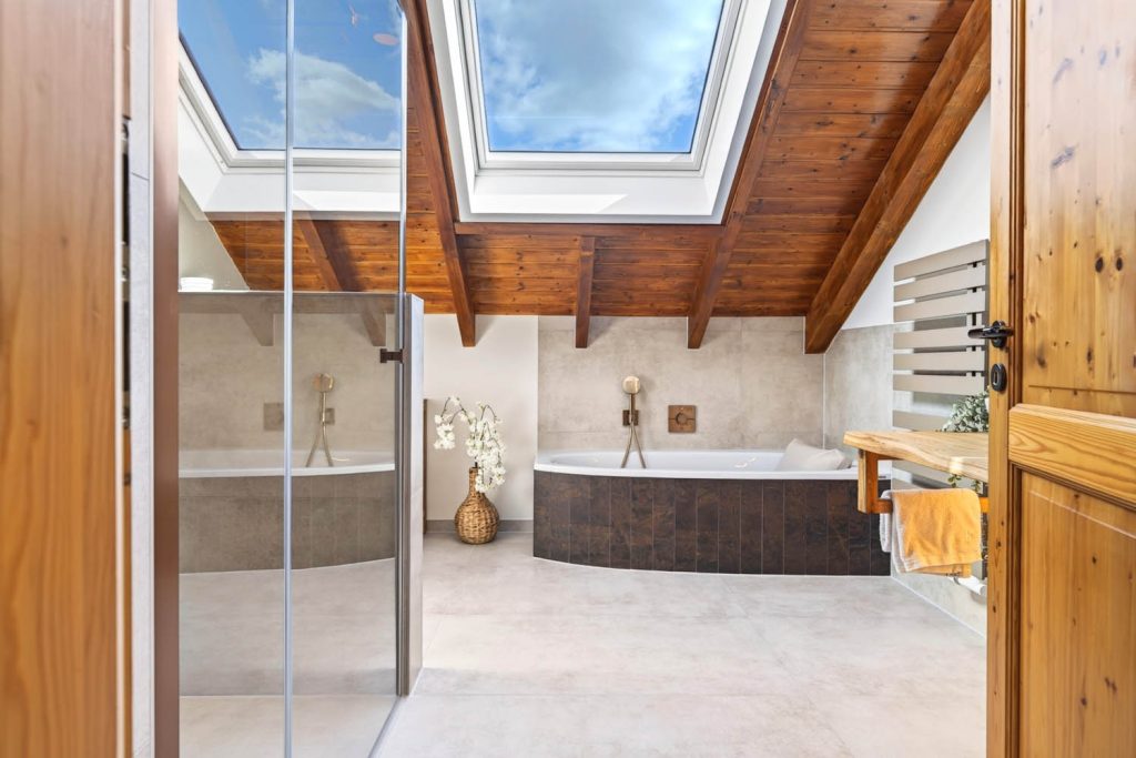 Blick durch die Holztür ins Dachbad mit sichtbaren Holzbalken, Eckbadewanne und Waschtisch
