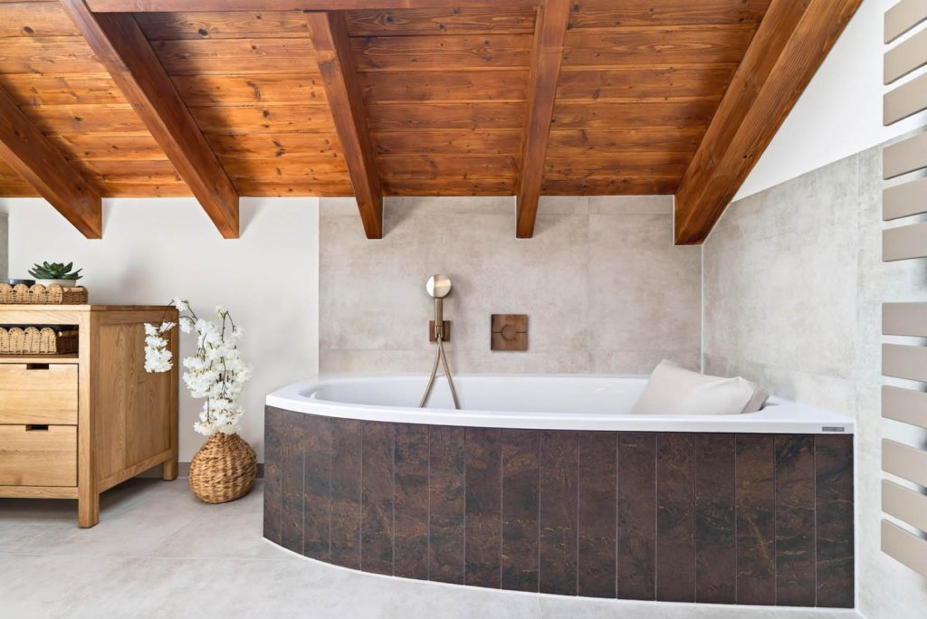 Eckbadewanne mit dunkler Verkleidung unter der Holzdecke, links die Holzkommode