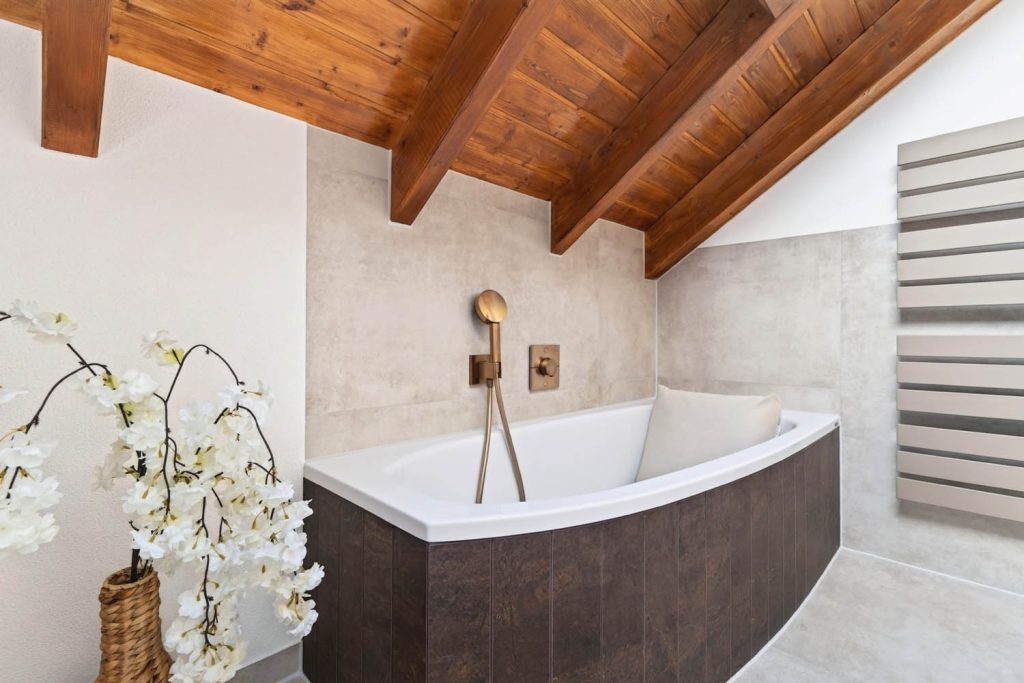 Eckbadewanne mit Armatur in Kupfer unter Holzbalken, im Vordergrund Blumendeko
