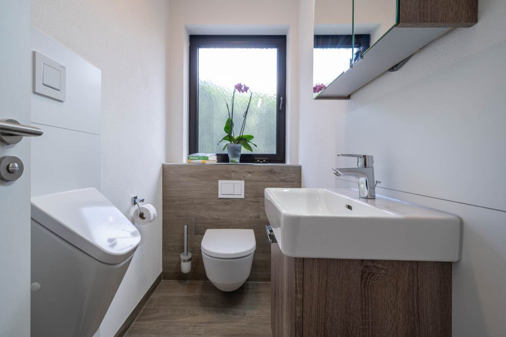 Gäste-WC mit Waschtisch, Wand-WC vor holzfarbener Vorwand und Fenster mit Orchidee, links angeschnitten ein Urinal