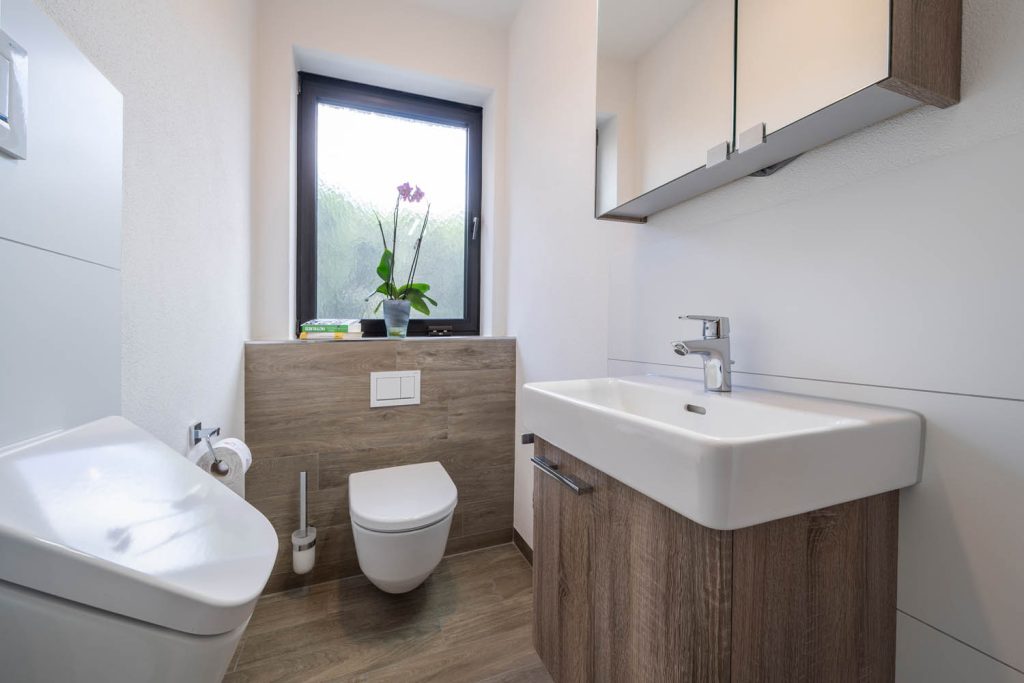 Waschbecken auf Unterschrank in Holzoptik, dahinter Wand-WC unter dem Fenster, vorne links ein Urinal