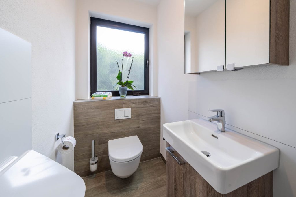 Gäste-WC mit Hängeschrank, Waschtisch und Wand-WC vor Vorwand in Holzoptik, links ein Urinal im Anschnitt