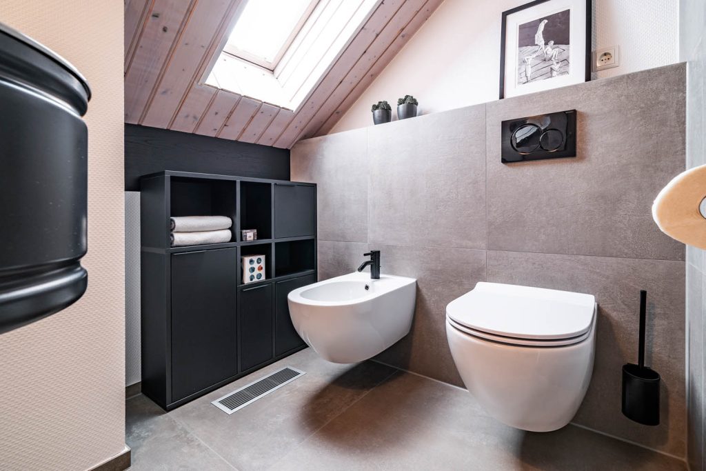 Bidet und Wand-WC vor großformatigen grauen Fliesen, links schwarzes Regal