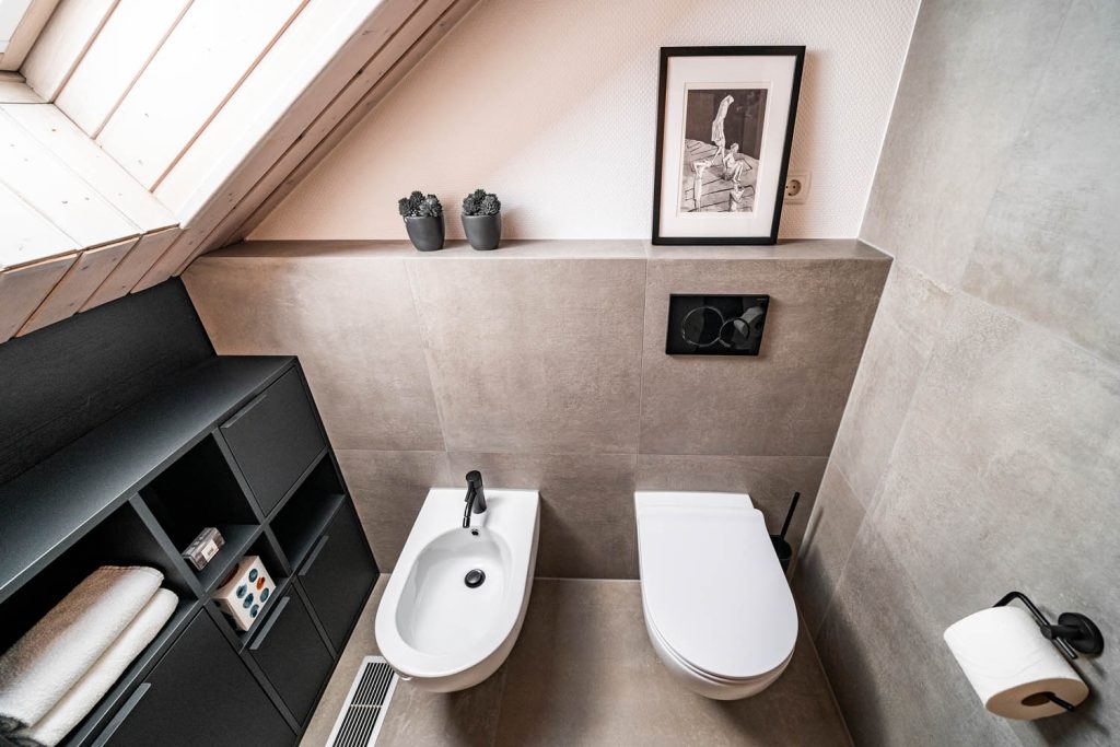Bidet und WC vor grauer Wand, links Regal mit Handtüchern