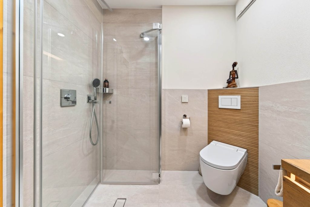 Eckdusche mit Glastüren neben dem Wand-WC vor heller Fliesenwand